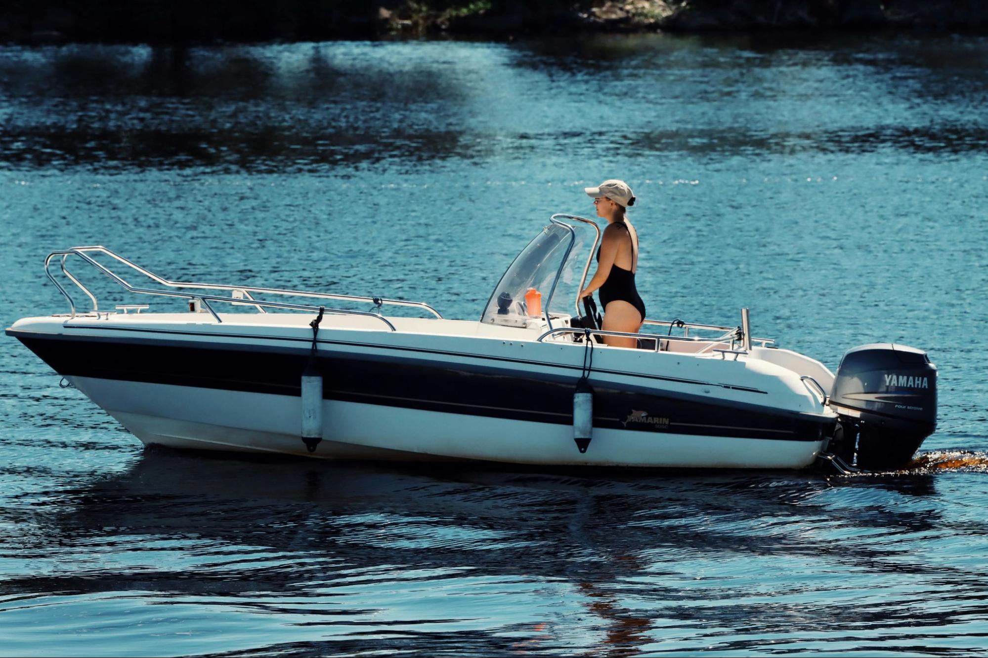 A person in a swimsuit and a baseball cap stands at the helm of a small motorboat on calm blue water.
