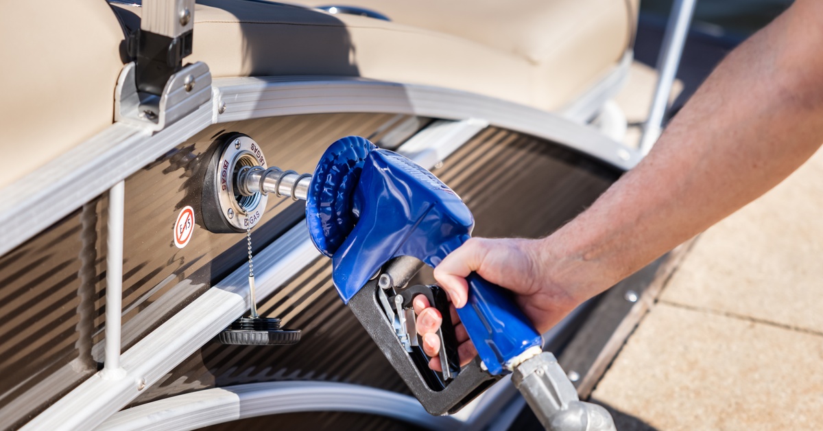 Comparing Electric vs. Gas Boating: What You Should Know A close-up of a man's hand refueling a boat at a marina. The blue gas pump nozzle is filling the pontoon party boat's fuel tank.