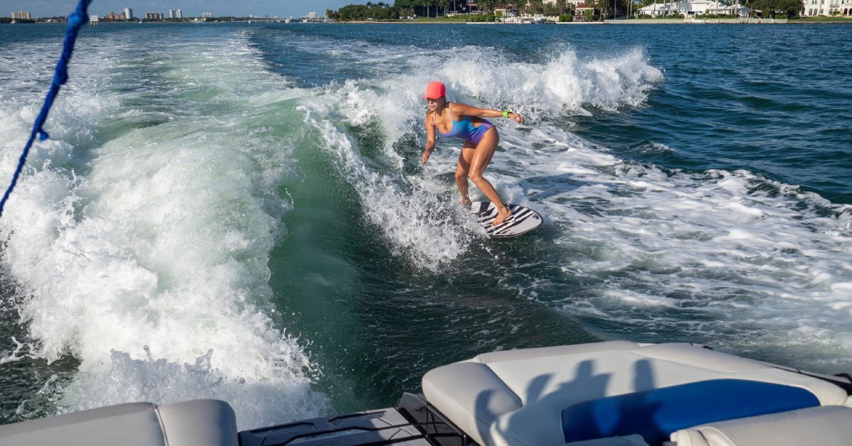 A woman wearing a red cap leans forward while wakesurfing behind a boat.