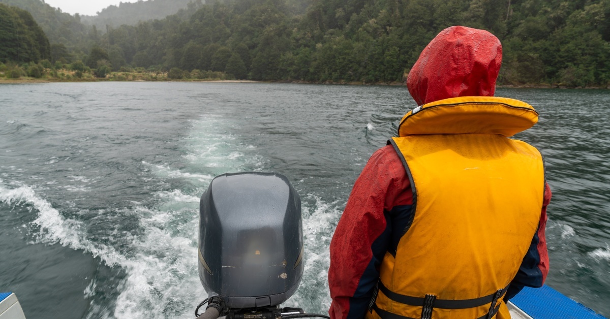 A man wearing a red rain jacket with a yellow life vest standing at the edge of his boat near the outboard motor.