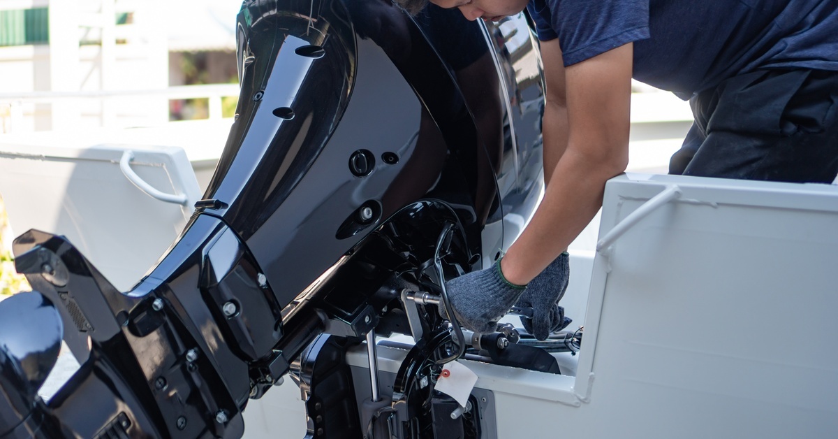 A mechanic wearing grey gloves leaning down to repair a boat’s outboard motor. The outboard motor is black and has a shiny finish.
