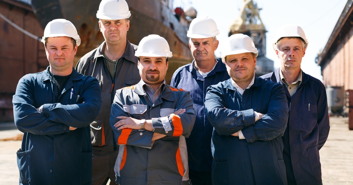 A group of men wearing dark blue and gray outfits with white hard hats, standing together in front of a ship at a shipyard. A group of men wearing dark blue and gray outfits with white hard hats, standing together in front of a ship at a shipyard.