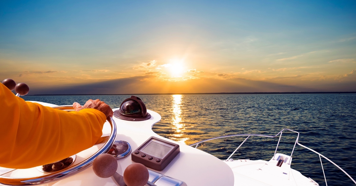What Makes the ePropulsion Spirit Electric Outboard Unique? The hand of a person wearing an orange jacket is on the steering wheel of a motorboat with the sun setting in front of him.