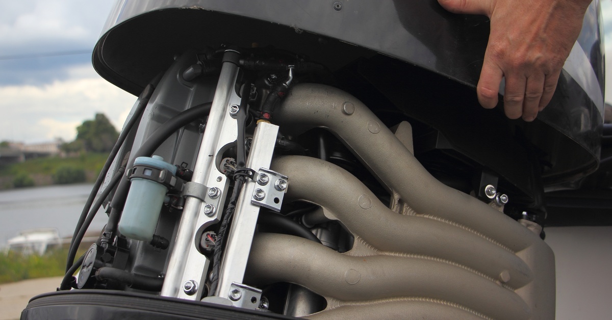 A person lifts the cover of an outboard motor, revealing the engine components and exhaust manifold near the water’s edge. A person lifts the cover of an outboard motor, revealing the engine components and exhaust manifold near the water’s edge.