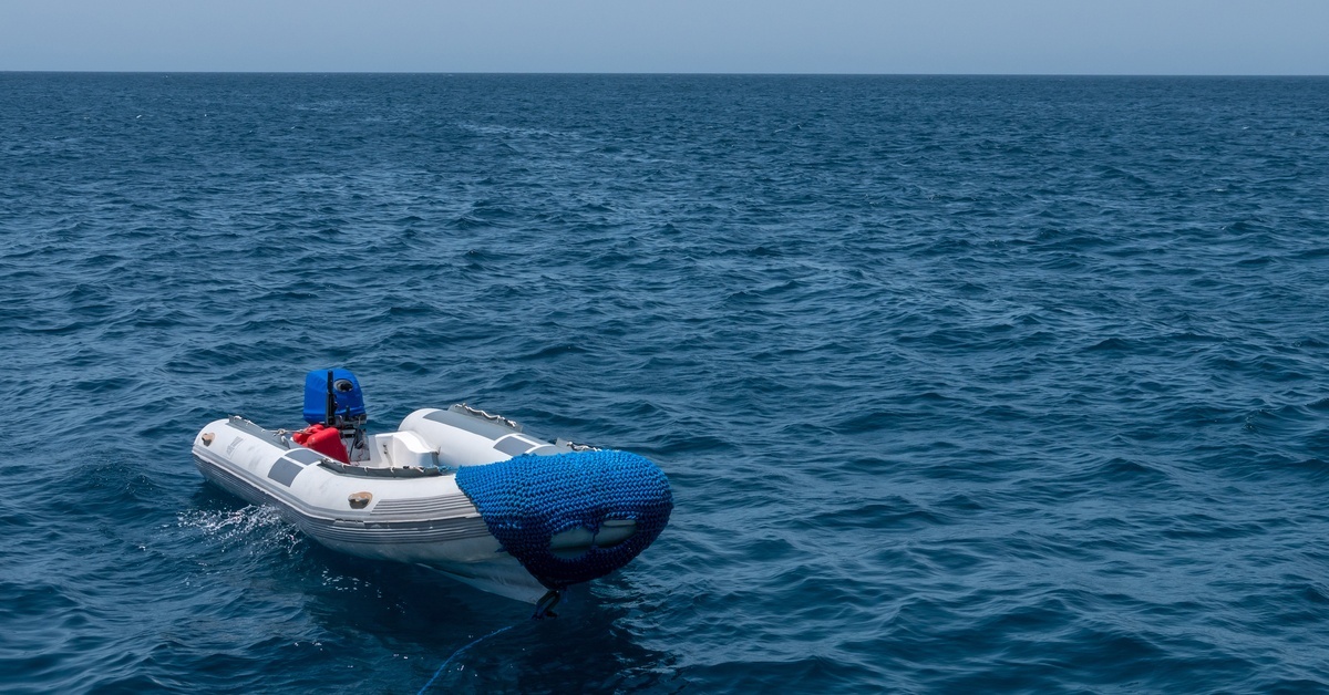 A white and gray RIB floating on open water. There is a blue motor on the back and a blue rope trailing along the front. A white and gray RIB floating on open water. There is a blue motor on the back and a blue rope trailing along the front.